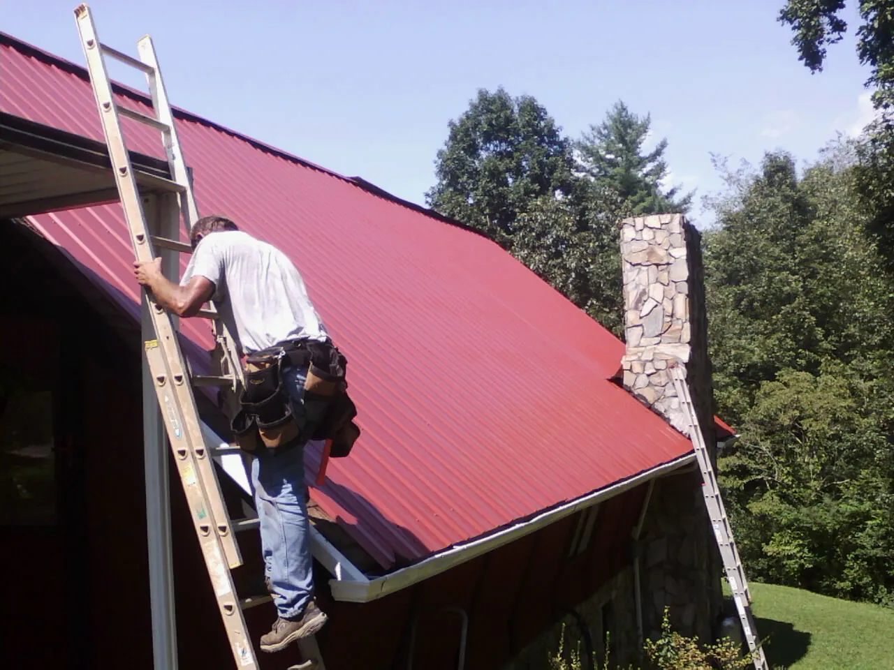 Roofer on a ladder beside a red metal roof and stone chimney — Soffit & Fascia Repair by Roofing Brothers in Huger